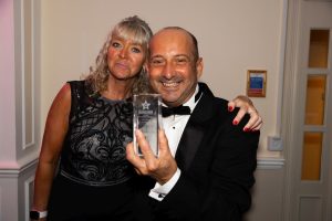 A man in a dinner jacket holds an award beside a smiling woman in a black dress at an indoor event.