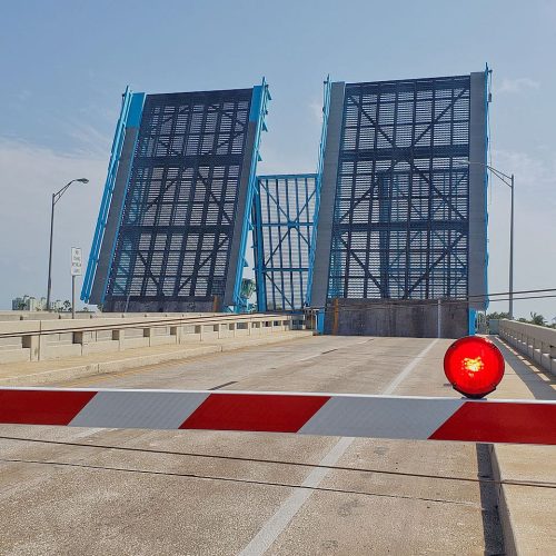 A drawbridge stands raised with a closed barrier and a glowing red signal light preventing vehicles from crossing.
