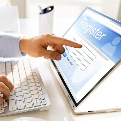 A person points at a computer screen showing an online registration form, with a keyboard and a small plant placed nearby.