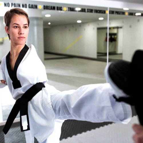 A woman wearing a martial arts uniform performs a high kick towards a target pad inside a gymnasium.