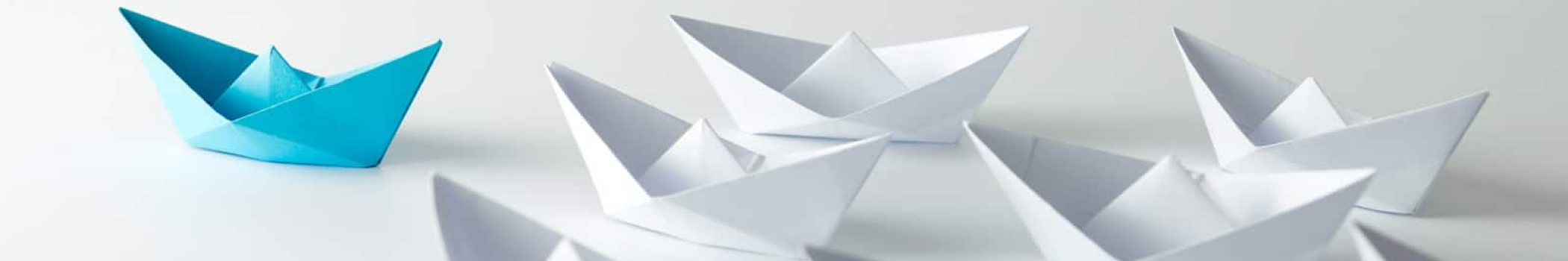 A single blue paper boat is positioned in front of a group of white paper boats.