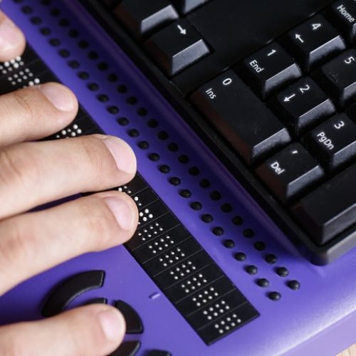 Hands reading a braille display positioned in front of a computer keyboard on a purple surface.