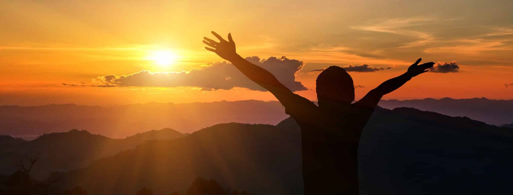 A person’s silhouette stands with arms raised, facing a bright sunrise above rolling mountain hills in the background.