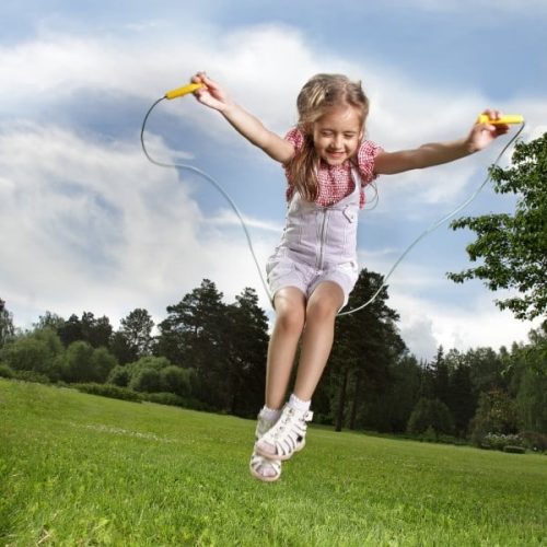 A girl in white dungarees smiles whilst skipping outside on green grass, with trees and a blue sky behind her.