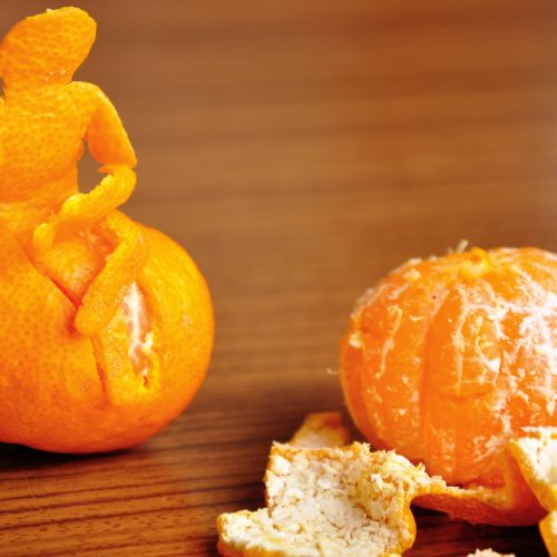 An orange with its peel arranged to resemble a seated person is beside a peeled orange and loose peel on a wooden table.
