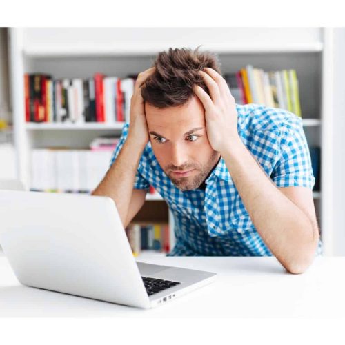 A man wearing a blue checked shirt appears stressed as he stares at a laptop in a bright room with bookcases.