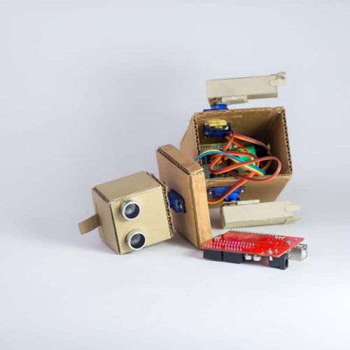 A small cardboard robot with exposed wires is lying on its side next to a red circuit board on a white background.