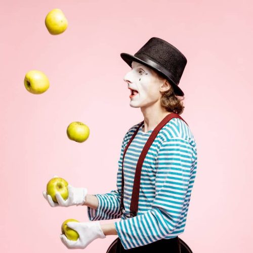 A mime wearing a striped shirt and hat juggles five green apples in front of a pink background.