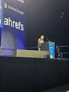 A person stands at a lectern onstage at a conference, with ahrefs and DemandSphere logos displayed on a screen behind them.