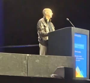 A man wearing a leather jacket stands at a lectern on stage, speaking into a microphone with a screen behind him.